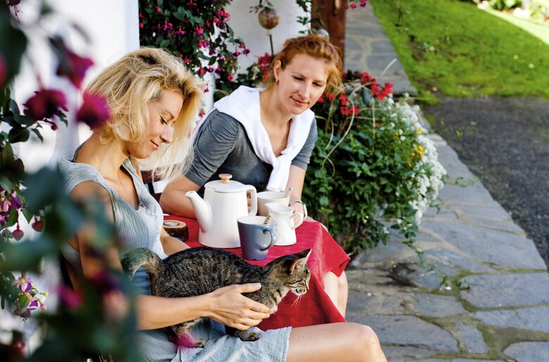 Frauen sitzen mit Katze auf der Hausbank, Plank | © Urlaub am Bauernhof / Kufferath Zwei Frauen sitzen gemütlich auf einer Hausbank mit einer Katze am Schoß | © Urlaub am Bauernhof / Kufferath
