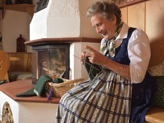 Oma im Dirndl strickt Socken am Kachelofen | © Urlaub am Bauernhof Oberösterreich / Harald Puchegger