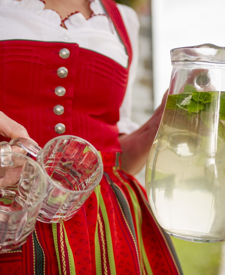 Bäuerin im roten Dirndl trägt einen Krug Kräutersirup mit frischer Melisse. | © Urlaub am Bauernhof Oberösterreich / Karin Lohberger