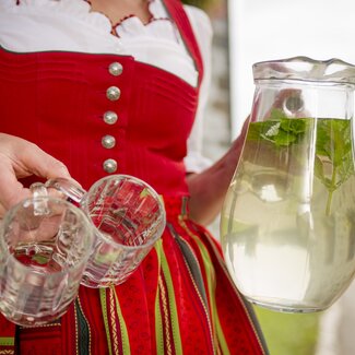 Bäuerin im roten Dirndl trägt einen Krug Kräutersirup mit frischer Melisse. | © Urlaub am Bauernhof Oberösterreich / Karin Lohberger