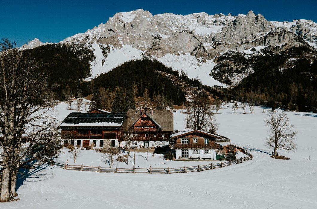 Außenansicht Hof | © Urlaub am Bauernhof / Daniel Gollner Traditionelles Bergdorf inmitten verschneiter Berglandschaft, umgeben von Wälder und einem Zaun. Mehrere Holzhäuser mit typischer Architektur sind zu sehen, einige mit Solarpanelen auf den Dächern. | © Urlaub am Bauernhof / Daniel Gollner