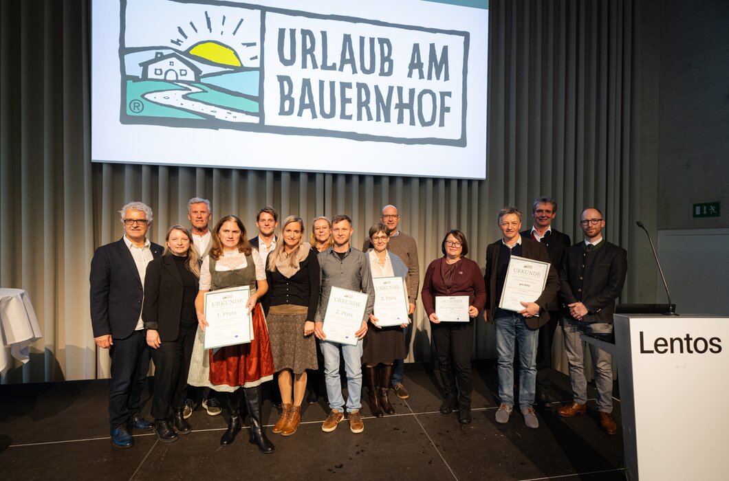 Alle Sieger des Innovationstages 2024  | © Urlaub am Bauernhof / Christian Lendl Eine Gruppe von Personen, einige in traditioneller Tracht, stehen auf einer Bühne und halten Urkunden. Im Hintergrund ist ein grafisches Logo eines landwirtschaftlichen Betriebs zu sehen. | © Urlaub am Bauernhof / Christian Lendl