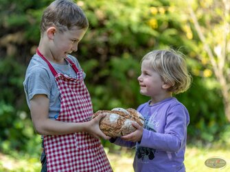 Zwei Kinder schauen sich an und geben sich einen Laib Brot | © Bio-Bergbauernhof Weger