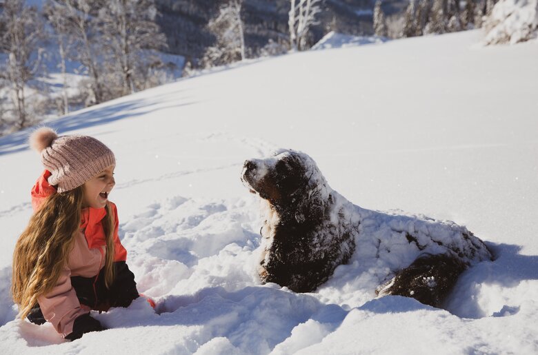 Mädchen mit Hund im Schnee | © Urlaub am Bauernhof / Pascal Baronit Ein Mädchen in roter Jacke sitzt im Schnee und spielt mit einem schwarzen Hund. Umgeben von schneebedeckten Bäumen in einer winterlichen Landschaft. | © Urlaub am Bauernhof / Pascal Baronit