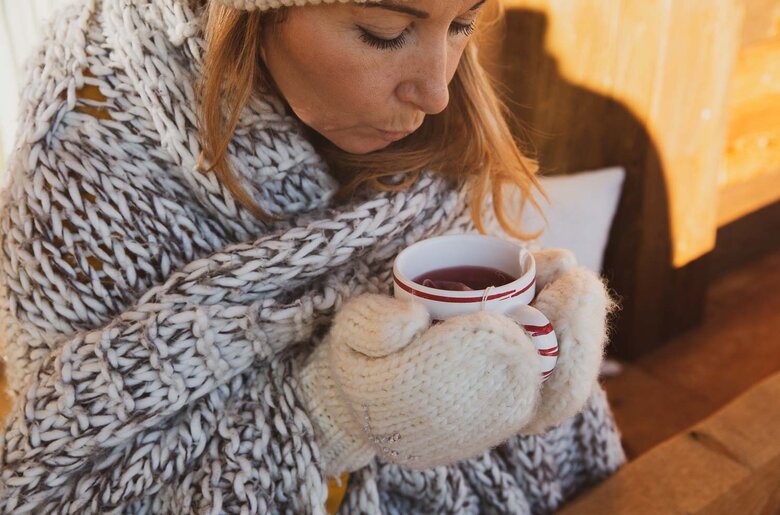 Frau bläst in Tasse Tee | © Urlaub am Bauernhof / Pascal Baronit In einem warmen Ambiente sitzt eine Person in einem gemütlichen, gestrickten Schal und genießt entspannt eine Tasse Tee. Die Stimmung ist gemütlich und einladend. | © Urlaub am Bauernhof / Pascal Baronit