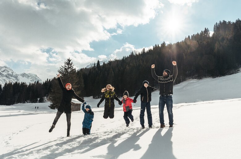 Familie springt im Schnee | © Urlaub am Bauernhof / Punkt und Komma Eine Gruppe von Menschen in Winterkleidung, die freudig mit erhobenen Armen posiert, umgeben von schneebedeckten Bergen und Wäldern unter wolkigem Himmel. | © Urlaub am Bauernhof / Punkt und Komma