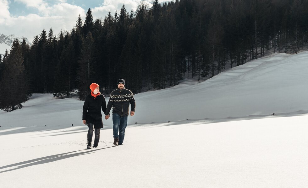 Spaziergang in schöner Schneelandschaft | © Urlaub am Bauernhof / Punkt und Komma Zwei Personen gehen Händchen haltend durch eine winterliche Landschaft mit Schneehügeln und verschneiter Berglandschaft im Hintergrund. | © Urlaub am Bauernhof / Punkt und Komma