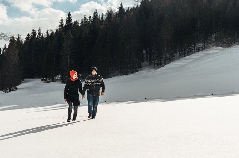 Spaziergang in schöner Schneelandschaft | © Urlaub am Bauernhof / Punkt und Komma Zwei Personen gehen Händchen haltend durch eine winterliche Landschaft mit Schneehügeln und verschneiter Berglandschaft im Hintergrund. | © Urlaub am Bauernhof / Punkt und Komma
