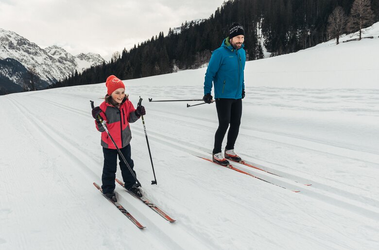 Langlaufen | © Urlaub am Bauernhof / Punkt und Komma Eine Person in Winterbekleidung, die Ski fährt, und eine weitere Person neben ihr, ebenfalls Ski fahrend, in einer schneebedeckten Winterlandschaft mit Bergen im Hintergrund. | © Urlaub am Bauernhof / Punkt und Komma