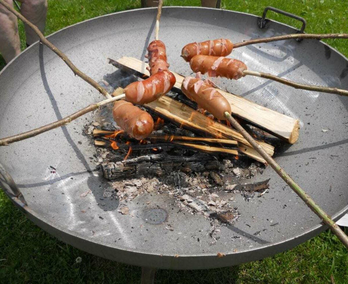 Knackwürste auf Grillstäben über einem offenen Feuer einer Grillschale.