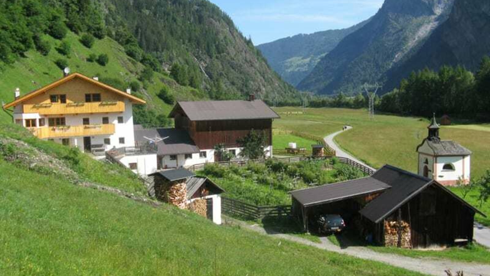 Blick von Süden (Hof im Gesamtblick): Räucherhaus, Heizlager mit Garage, hofeigene Kapelle mit Wohnhaus und Wirtschaftsgebäude; Blick über Wiesen zum Umhausener Talabschluss - dahinter Schi und Wandergebiet Hochötz